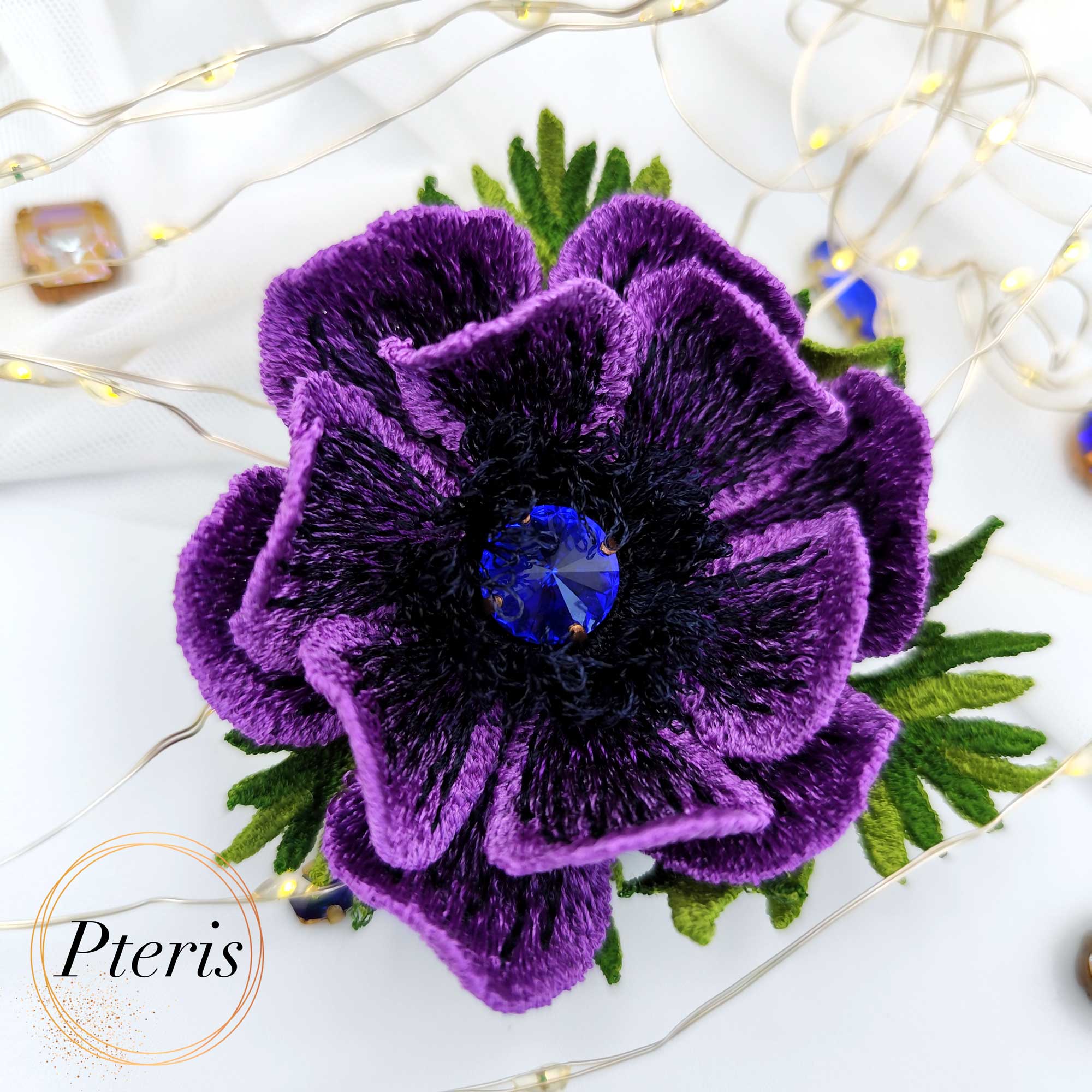 D Anemone Embroidery Design for FSL brooch featuring deep purple petals, a black textured center with a blue crystal, and green leaves.