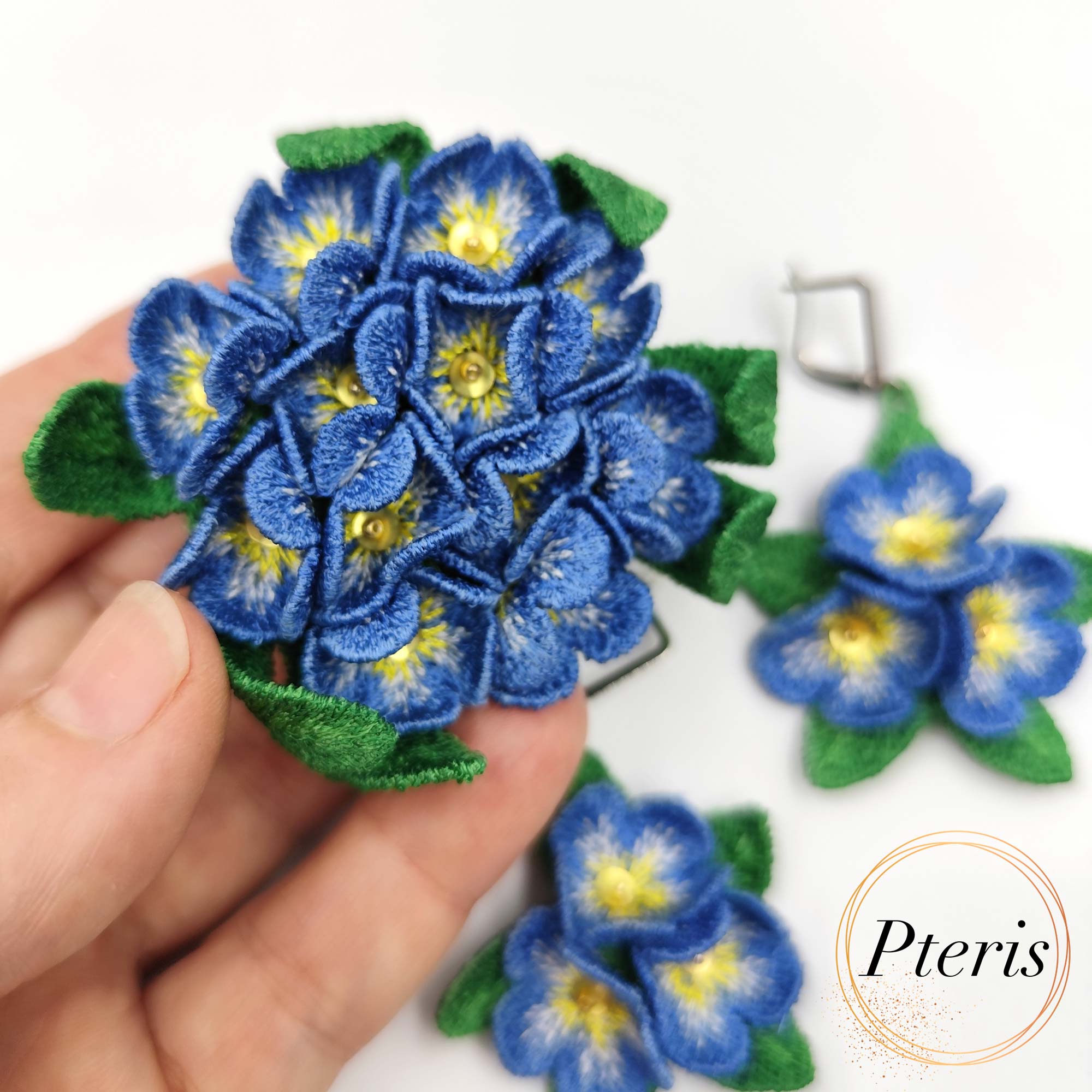 3D Forget-Me-Not Embroidery Design for FSL jewelry featuring blue flowers with yellow centers and green leaves on silver hooks.