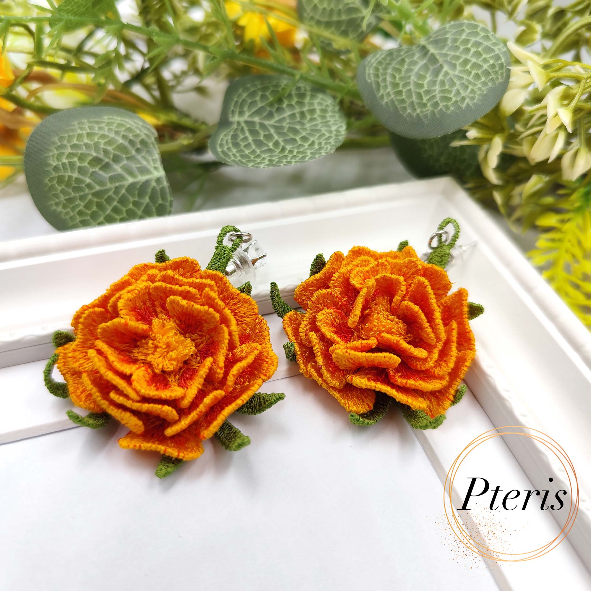 3D Marigold Embroidery Design for FSL jewelry featuring bright orange ruffled petals and green textured leaves on silver earring studs.