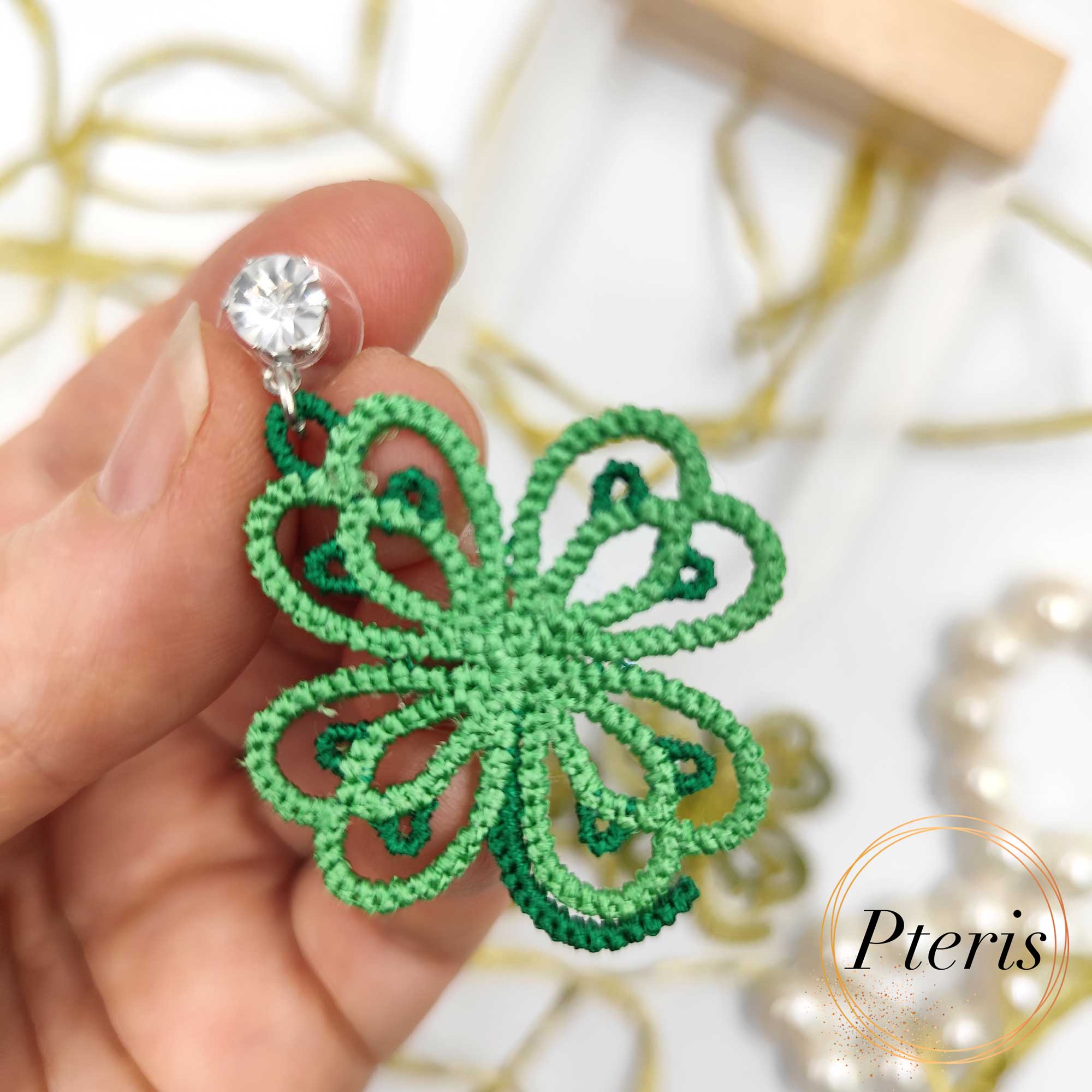 FSL Clover Earrings Embroidery Design featuring a pair of green tatting-style lace clover earrings with crystal studs, displayed among pearls and gold ribbons.