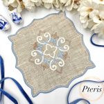 ITH Ornament Cocktail Napkin embroidery design showing a beige linen napkin with a blue and white central scroll ornament and blue satin-stitched edges, styled with white flowers and a blue velvet ribbon.