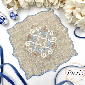 ITH Ornament Cocktail Napkin embroidery design showing a beige linen napkin with a blue and white central scroll ornament and blue satin-stitched edges, styled with white flowers and a blue velvet ribbon.