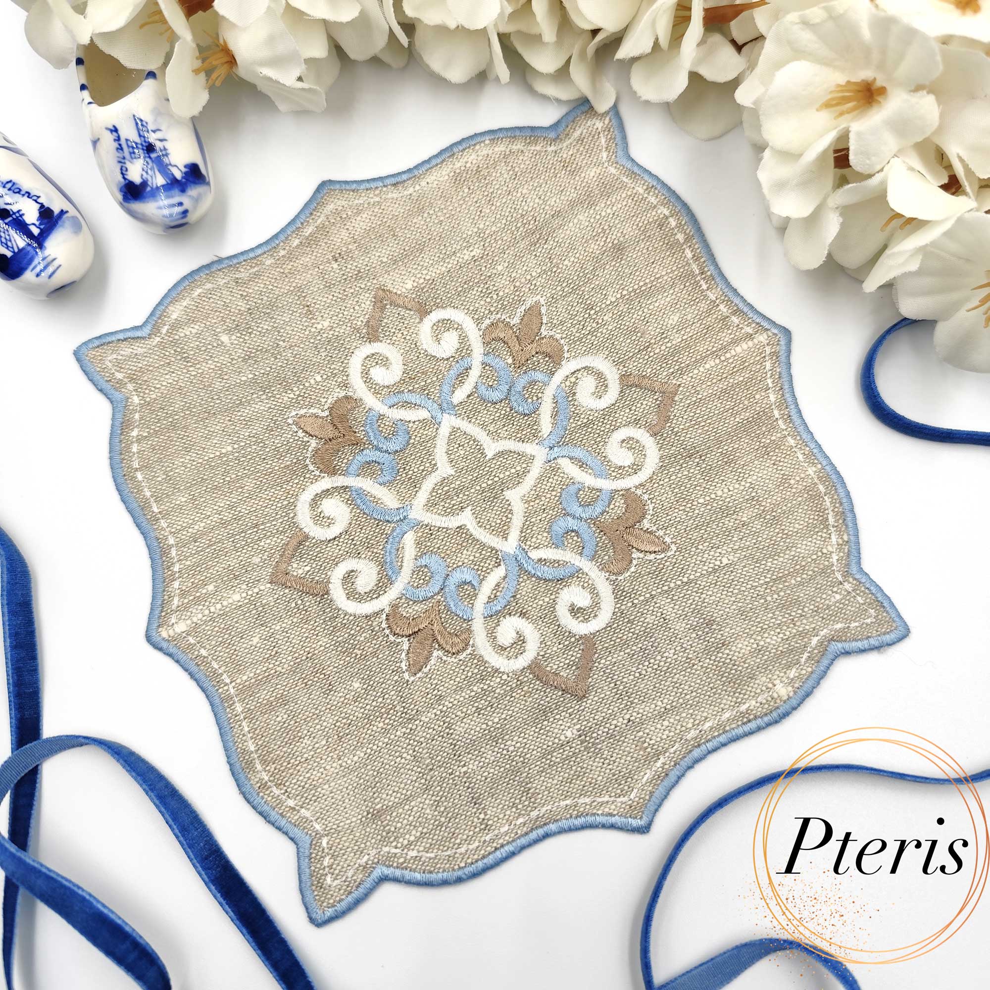 ITH Ornament Cocktail Napkin embroidery design showing a beige linen napkin with a blue and white central scroll ornament and blue satin-stitched edges, styled with white flowers and a blue velvet ribbon.