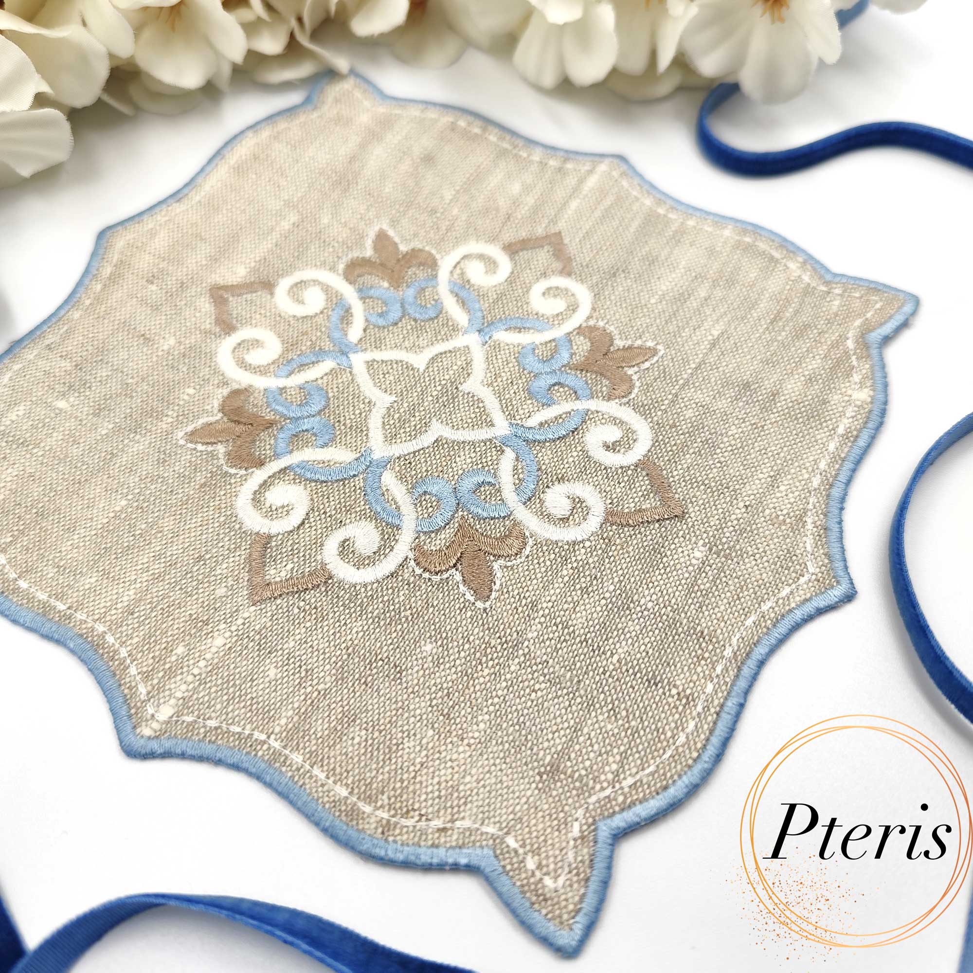 ITH Ornament Cocktail Napkin embroidery design showing a beige linen napkin with a blue and white central scroll ornament and blue satin-stitched edges, styled with white flowers and a blue velvet ribbon.