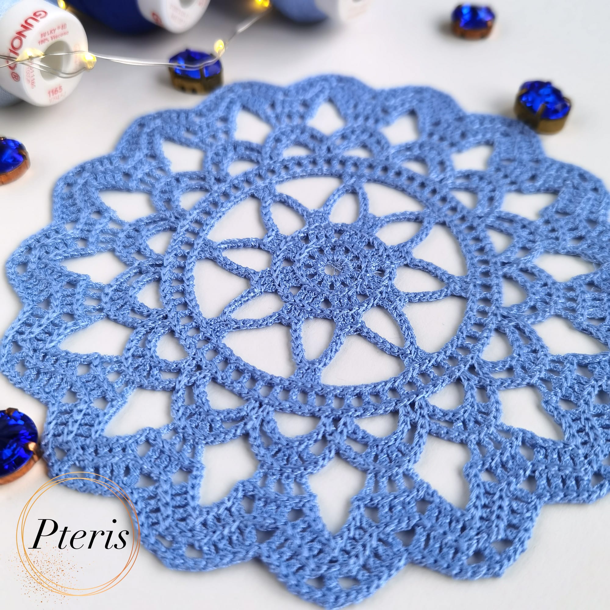 FSL Crochet Coaster Embroidery Design featuring two blue freestanding lace doilies with an intricate floral-web pattern, styled with blue thread spools and fairy lights.