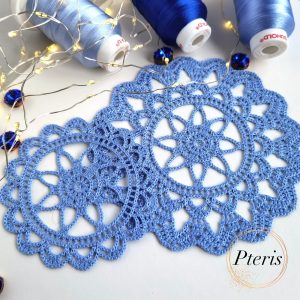 FSL Crochet Coaster Embroidery Design featuring two blue freestanding lace doilies with an intricate floral-web pattern, styled with blue thread spools and fairy lights.