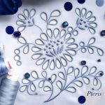 Detailed Richelieu cutwork machine embroidery design featuring blue floral motifs and openwork on white fabric, styled with blue thread spools and sparkling crystals.