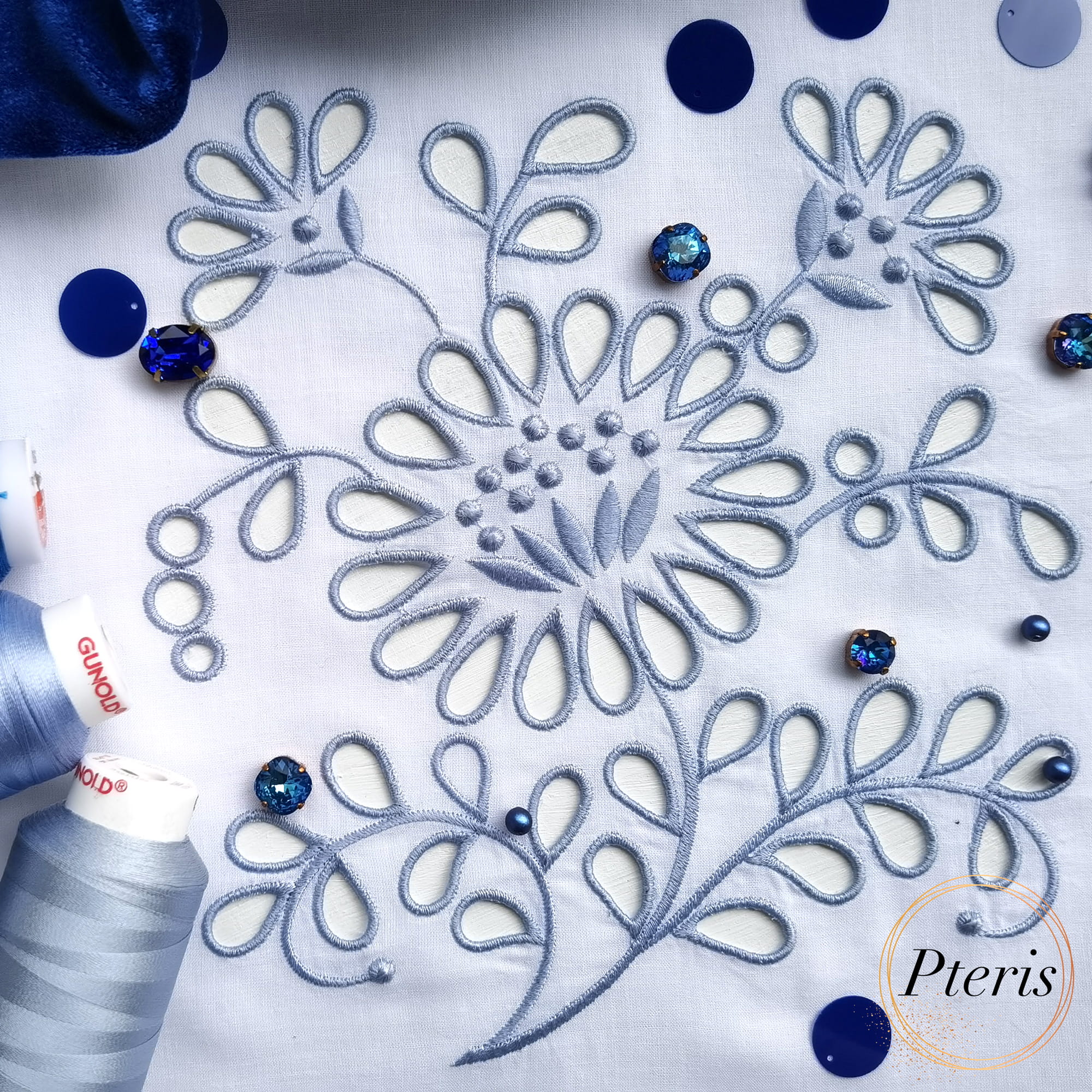 Detailed Richelieu cutwork machine embroidery design featuring blue floral motifs and openwork on white fabric, styled with blue thread spools and sparkling crystals.