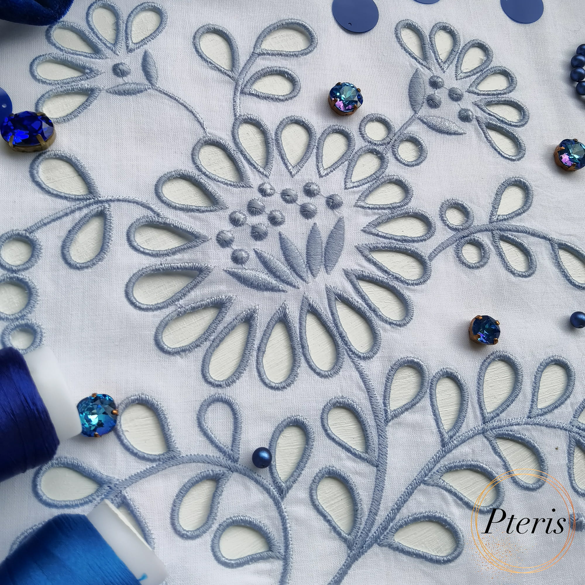 Detailed Richelieu cutwork machine embroidery design featuring blue floral motifs and openwork on white fabric, styled with blue thread spools and sparkling crystals.
