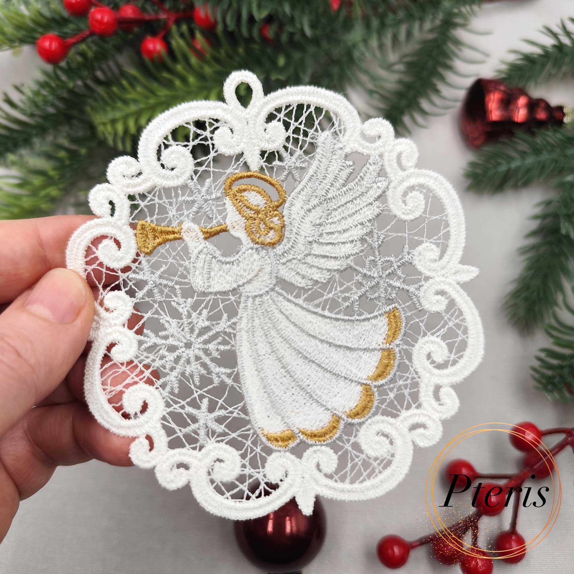 FSL Trumpeting Angel Suncatcher embroidery design featuring two white and gold freestanding lace medallions with angels, styled on a grey background with pine branches and red Christmas ornaments.