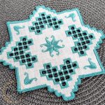 Blue Hardanger machine embroidery design featuring a white square cocktail napkin with intricate blue lace cutwork and star motifs, styled with silver ornaments and pine branches.