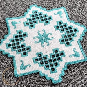Blue Hardanger machine embroidery design featuring a white square cocktail napkin with intricate blue lace cutwork and star motifs, styled with silver ornaments and pine branches.