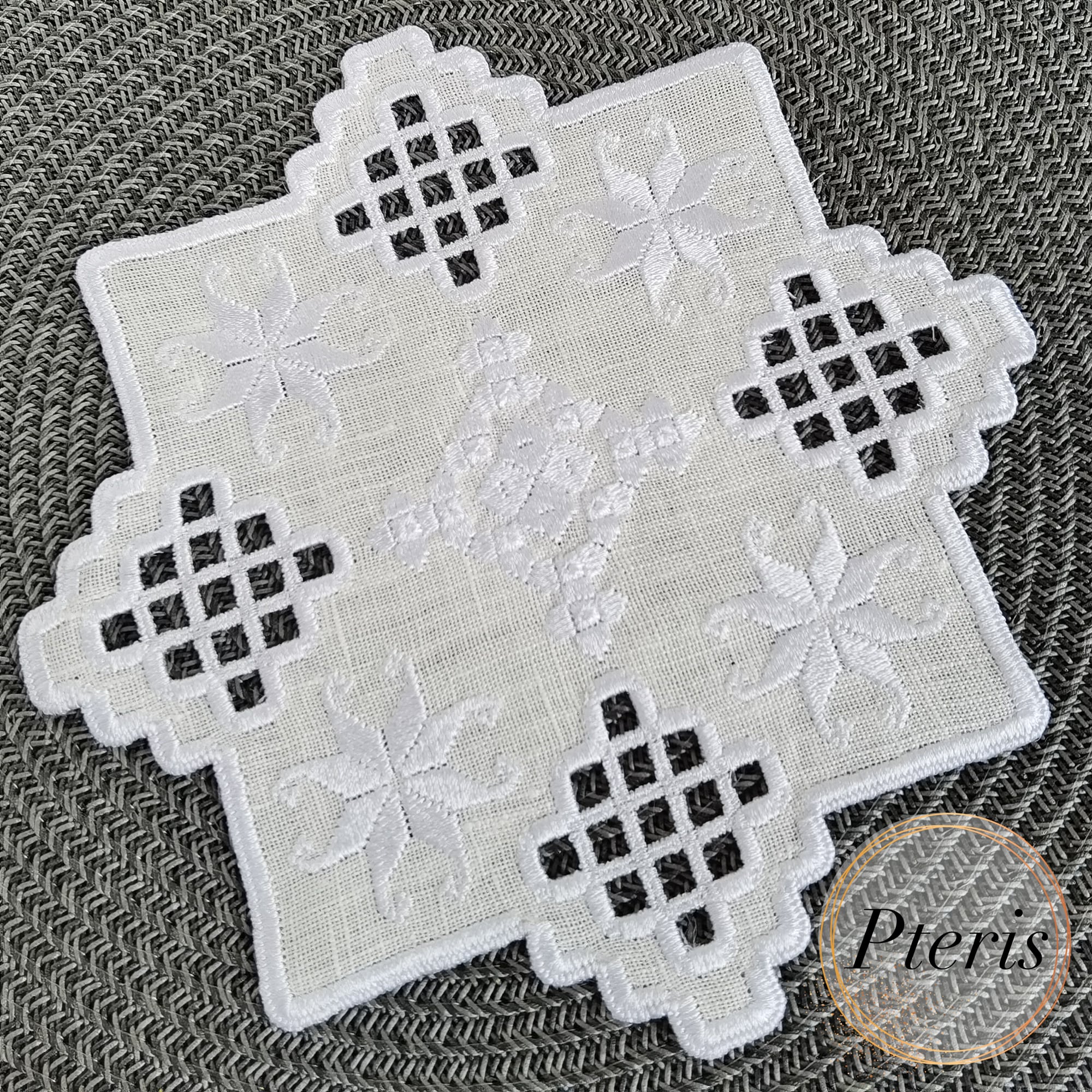 Hardanger machine embroidery design featuring a white square cocktail napkin with intricate lace cutouts and star motifs, styled on a grey background with pine branches and a star-shaped ornament.