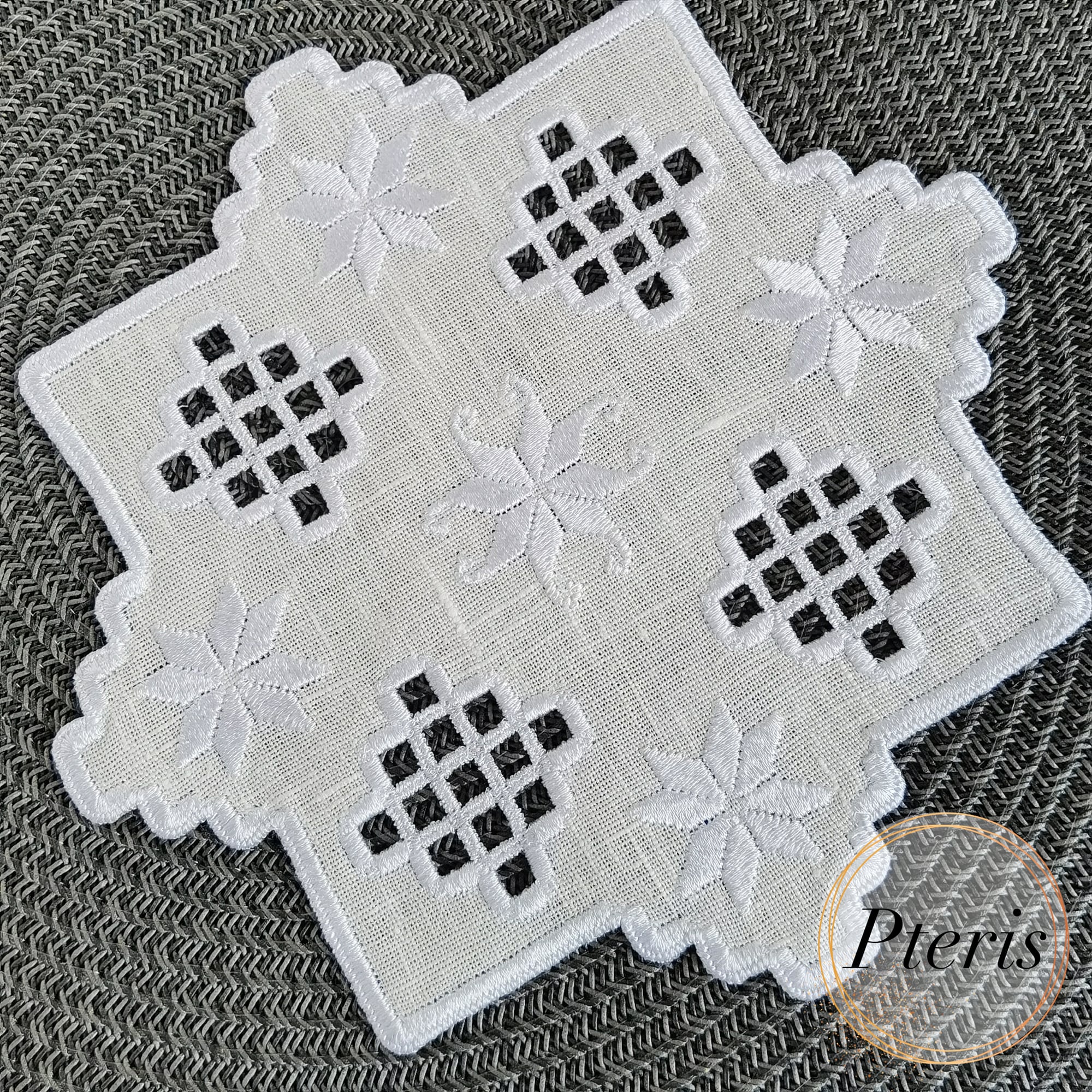 Hardanger machine embroidery design of a white linen cocktail napkin with pastel green and peach lace cutwork and floral stars, featuring a variegated scalloped edge on a denim background.