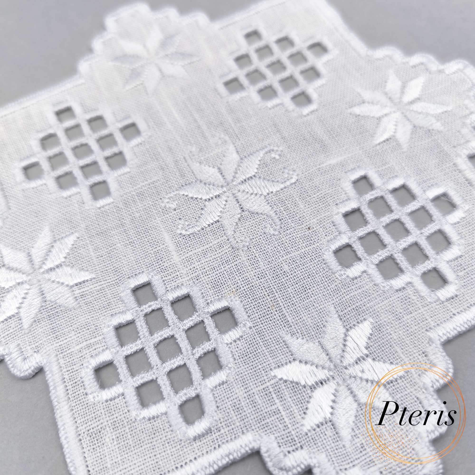 Hardanger machine embroidery design of a white linen cocktail napkin with pastel green and peach lace cutwork and floral stars, featuring a variegated scalloped edge on a denim background.