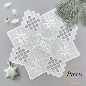 Hardanger machine embroidery design featuring a white square cocktail napkin with intricate lace cutouts and star motifs, styled on a grey background with pine branches and a star-shaped ornament.
