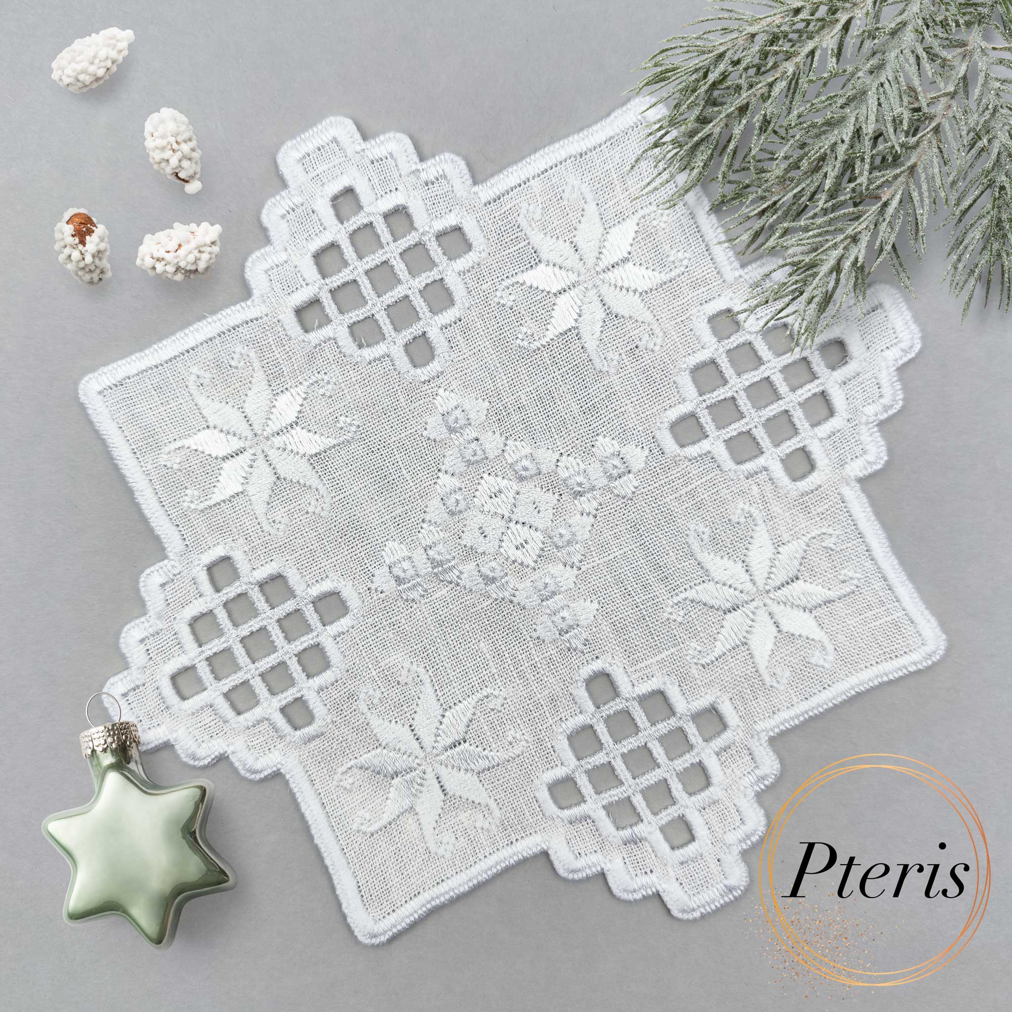 Hardanger machine embroidery design featuring a white square cocktail napkin with intricate lace cutouts and star motifs, styled on a grey background with pine branches and a star-shaped ornament.