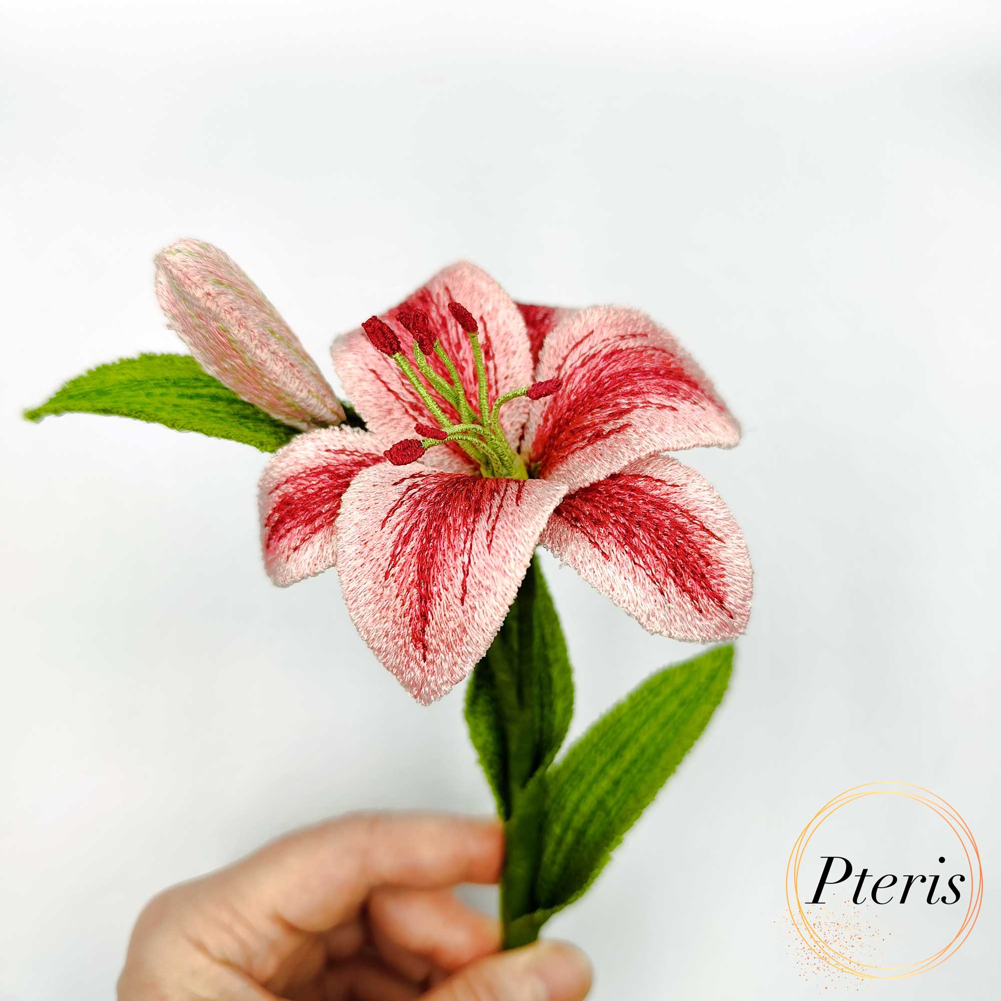 3D Lily machine embroidery design featuring a hand holding a highly detailed freestanding pink and red embroidered lily flower with a realistic green stem, bud, and textured leaves.