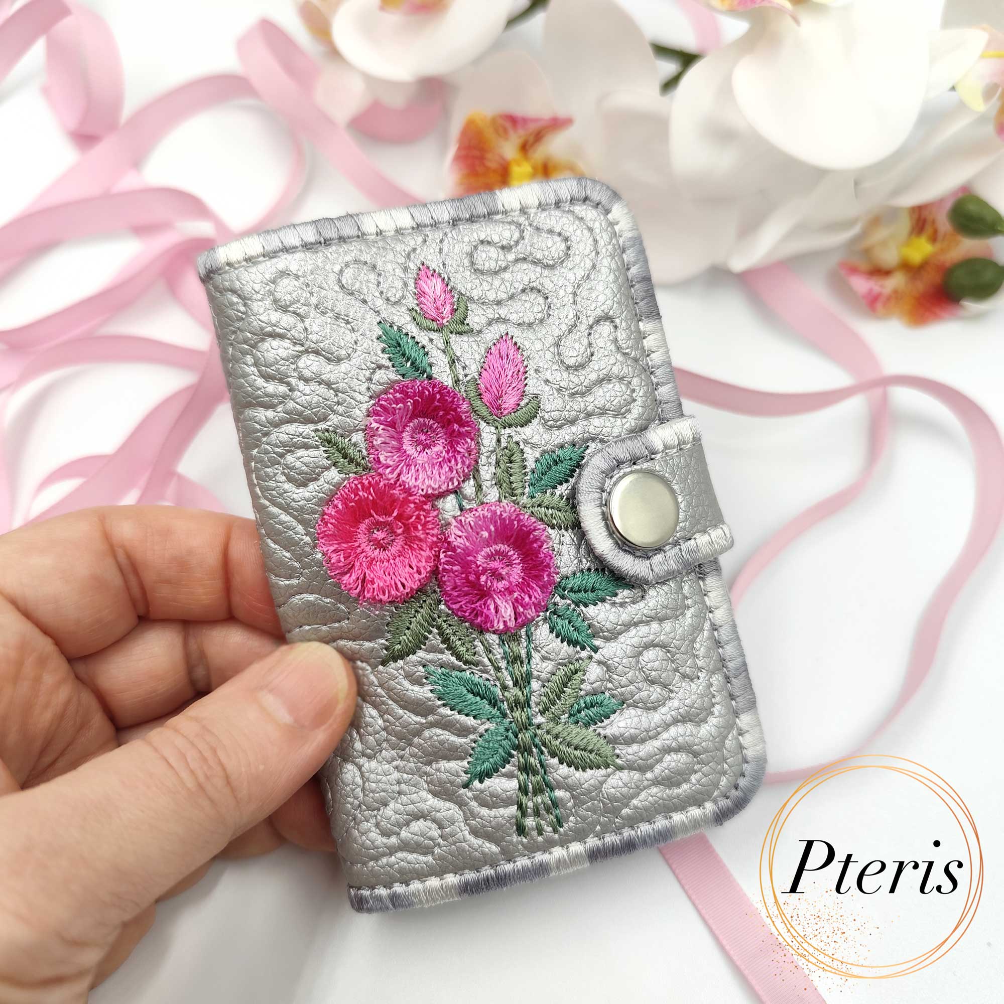 ITH Quilted Card Holder embroidery design featuring a metallic silver bifold wallet with vibrant pink fluffy flower embroidery and a snap closure, held in a hand.