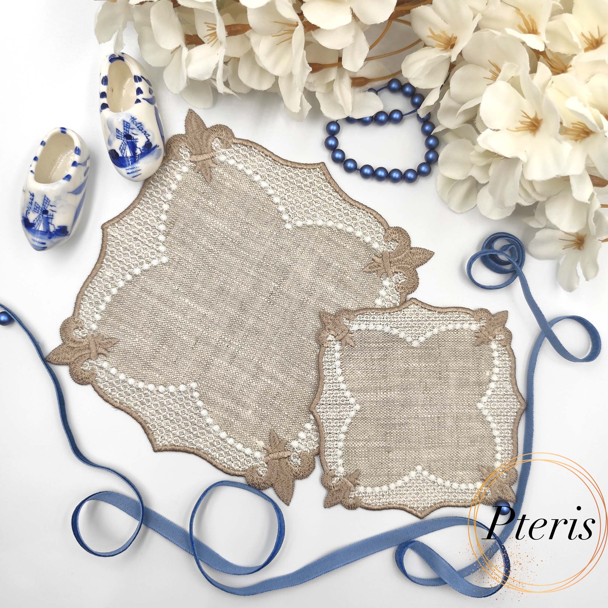 ITH Lace Cocktail Napkin embroidery design showing two different sized linen napkins with beige lace borders, styled with white flowers, blue beads, and miniature blue and white clogs.