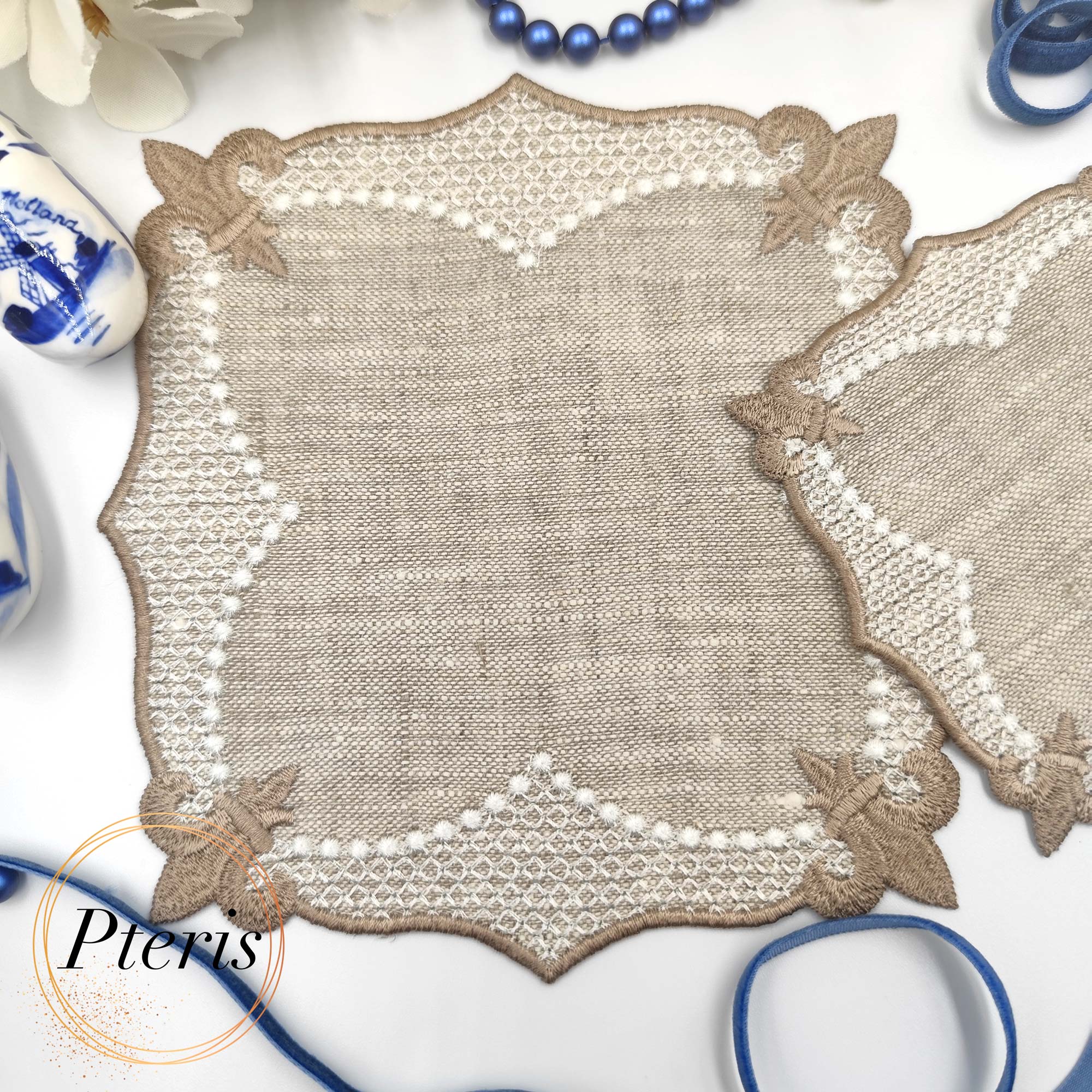 ITH Lace Cocktail Napkin embroidery design showing two different sized linen napkins with beige lace borders, styled with white flowers, blue beads, and miniature blue and white clogs.