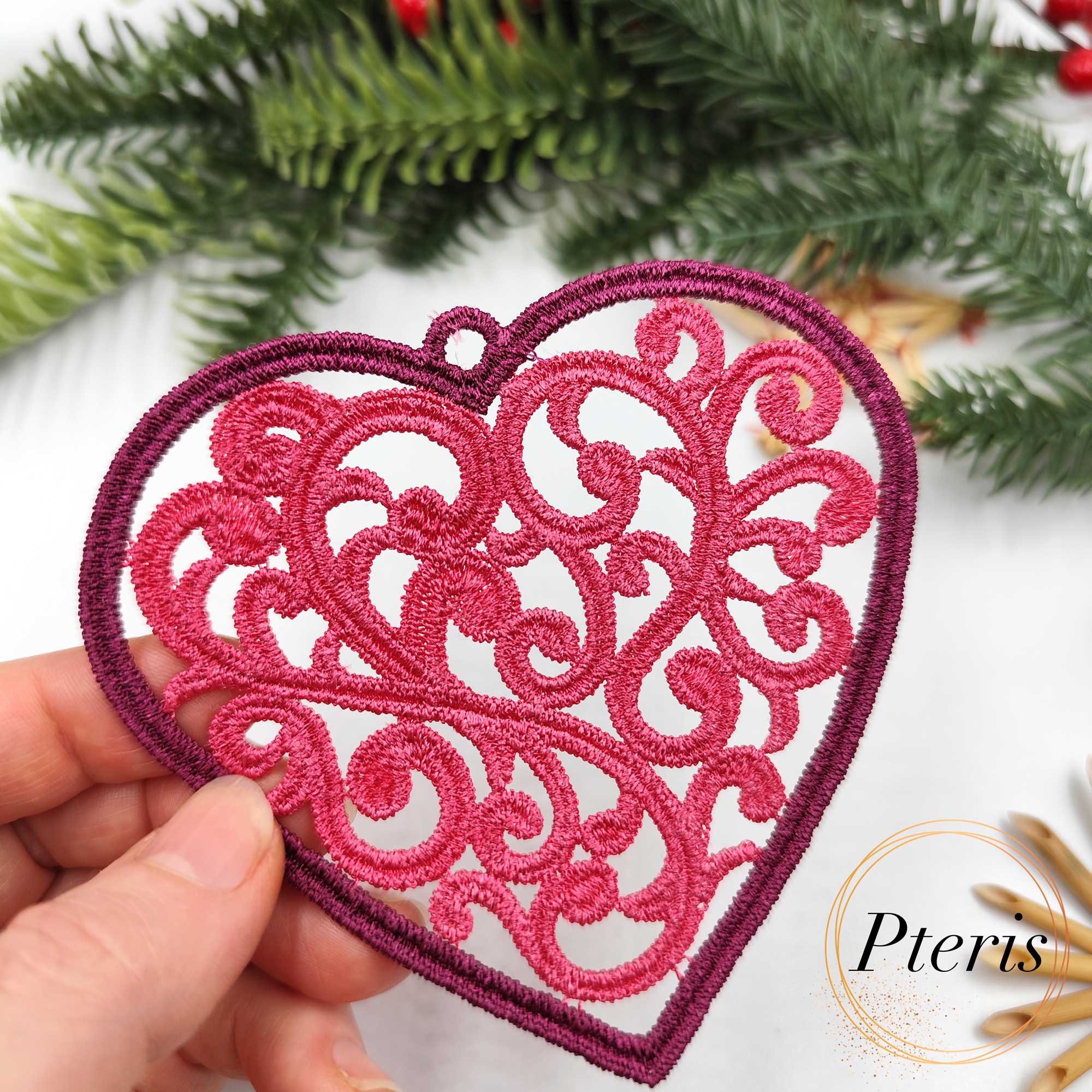 FSL Filigree Heart Suncatcher embroidery design featuring a pink freestanding lace heart with intricate scrolls, styled on a white background with pine branches and Christmas ornaments.