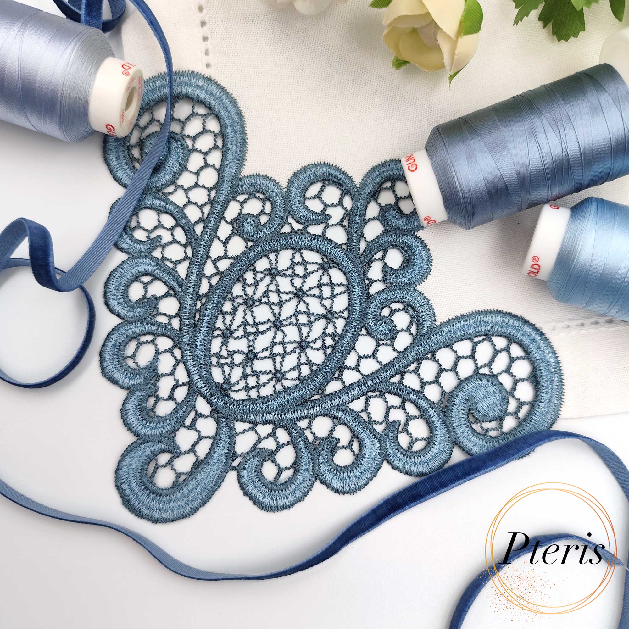 FSL Lace Napkin Corner embroidery design featuring intricate dusty blue freestanding lace stitched onto a white linen napkin, styled with matching blue thread spools and velvet ribbon.