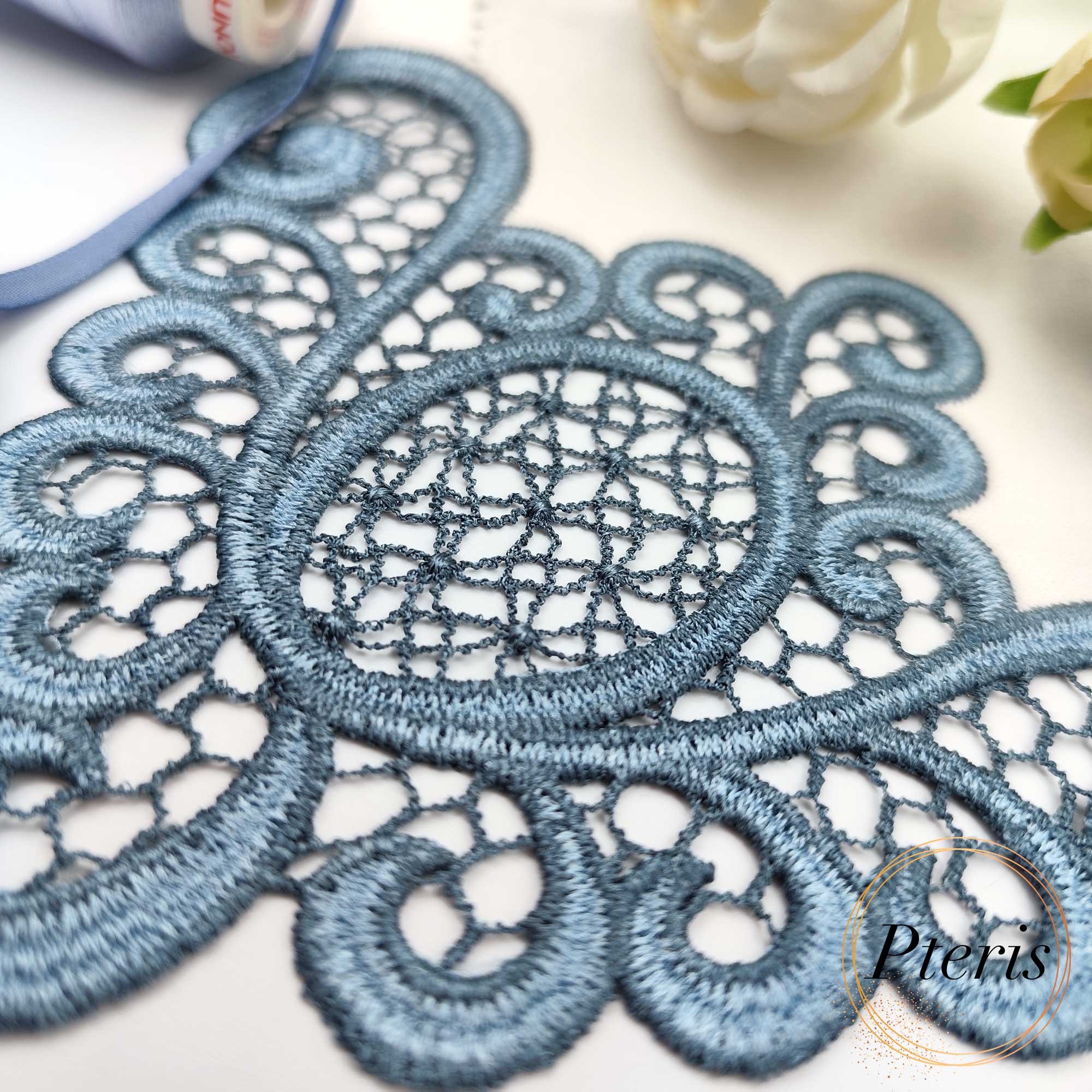 FSL Lace Napkin Corner embroidery design featuring intricate dusty blue freestanding lace stitched onto a white linen napkin, styled with matching blue thread spools and velvet ribbon.
