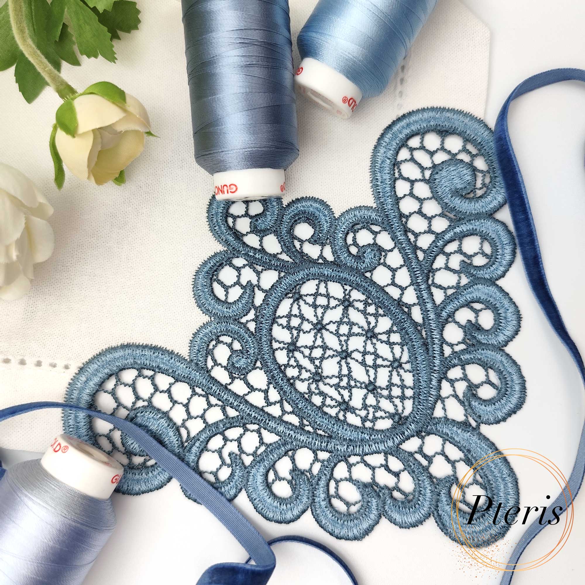 FSL Lace Napkin Corner embroidery design featuring intricate dusty blue freestanding lace stitched onto a white linen napkin, styled with matching blue thread spools and velvet ribbon.