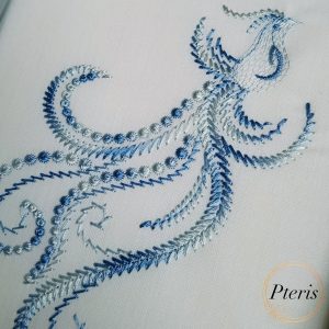 Bird with a flying feather machine embroidery design showing a gracefully stitched stylized bird trailing an elegant, flowing ornamental line-work feather motif, displayed flat on light blue fabric.