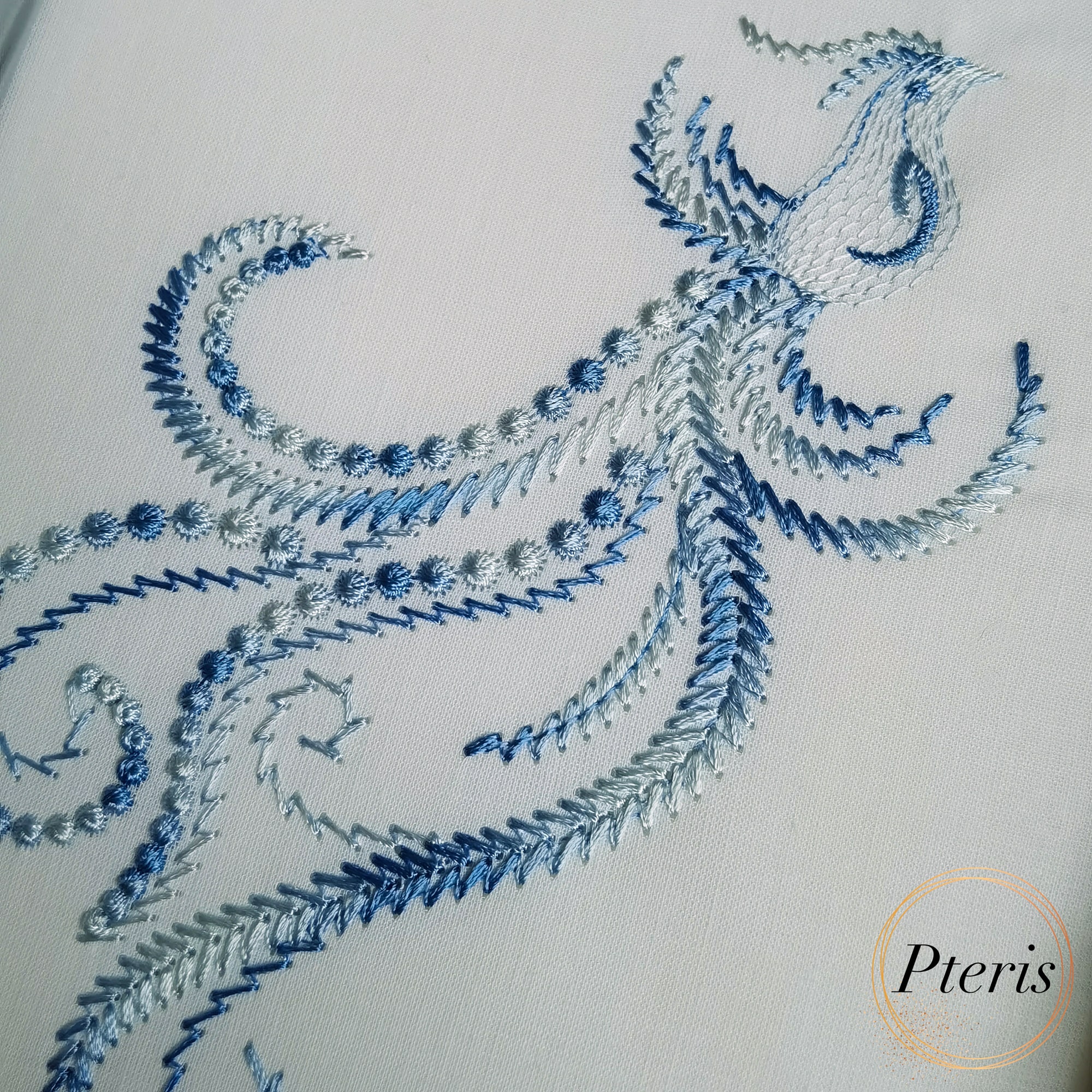 Bird with a flying feather machine embroidery design showing a gracefully stitched stylized bird trailing an elegant, flowing ornamental line-work feather motif, displayed flat on light blue fabric.