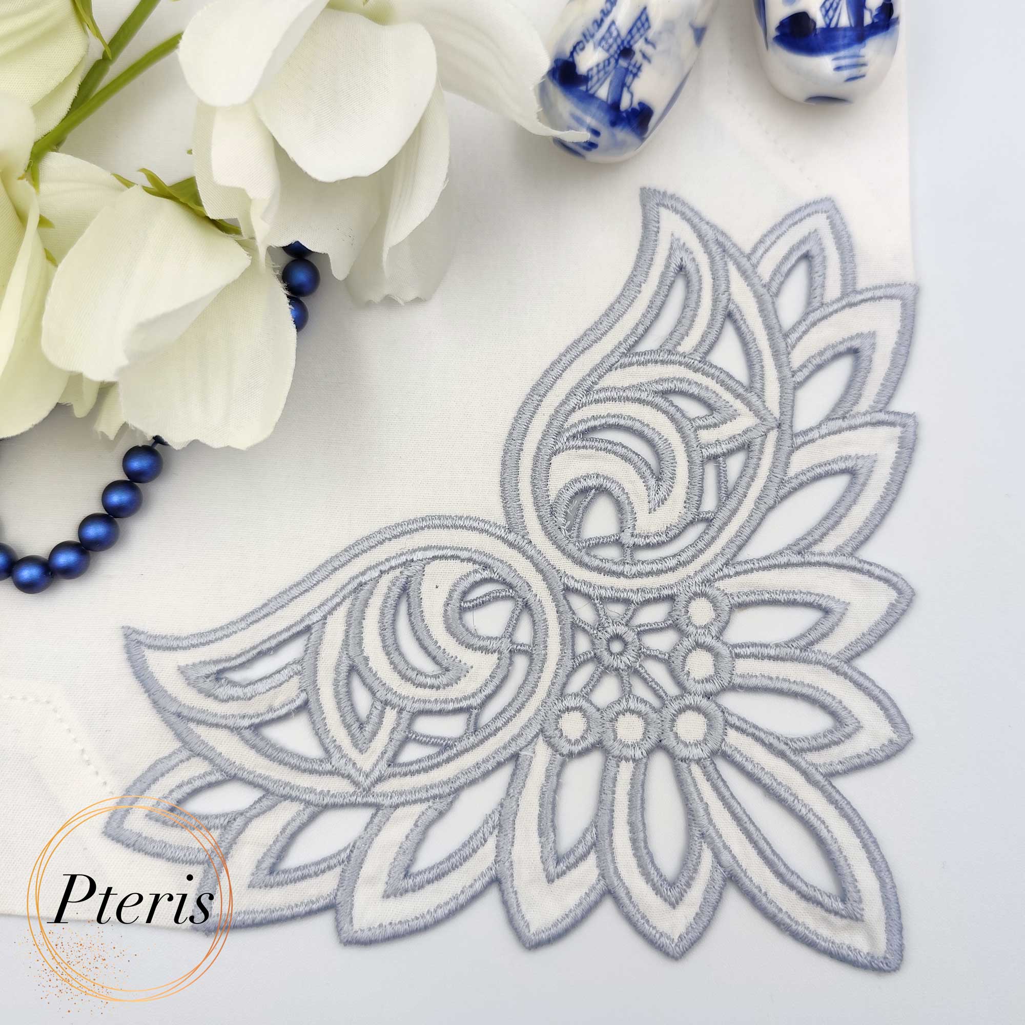 Richelieu cutwork machine embroidery design showcasing an elegant grey-blue floral corner pattern stitched onto a white fabric napkin, styled flat with white blossoms and blue beads.