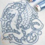 Regal Gryphon machine embroidery design featuring a close-up of an intricate mythical gryphon stitched in light blue-grey thread on white linen fabric, showcasing elegant Richelieu cutwork lace windows.