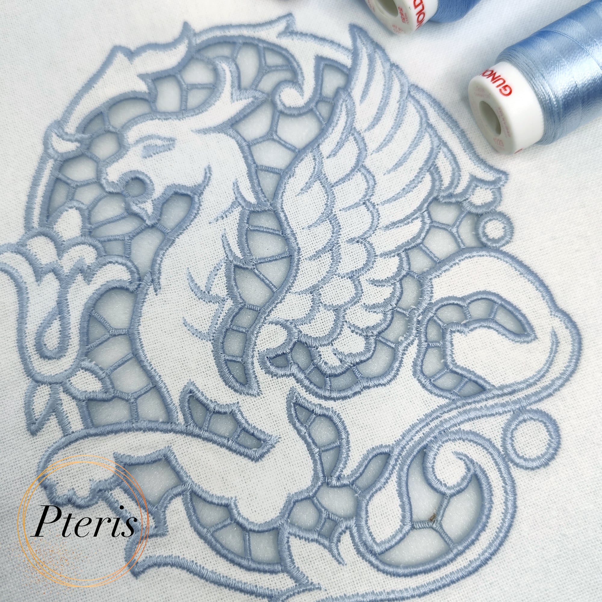 Regal Gryphon machine embroidery design featuring a close-up of an intricate mythical gryphon stitched in light blue-grey thread on white linen fabric, showcasing elegant Richelieu cutwork lace windows.