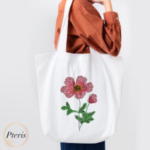 Flower Machine Embroidery Design (3).jpg showing a graceful sketch-style pink flower with a textured yellow-and-black center, green leaves, and a small flower bud, embroidered beautifully onto a white canvas tote bag held by a person.