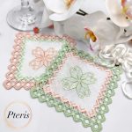 Square Hardanger cocktail napkin machine embroidery design displaying two linen napkins with a light green and peach embroidered geometric cutwork lace frame, styled flat next to white orchid blossoms.