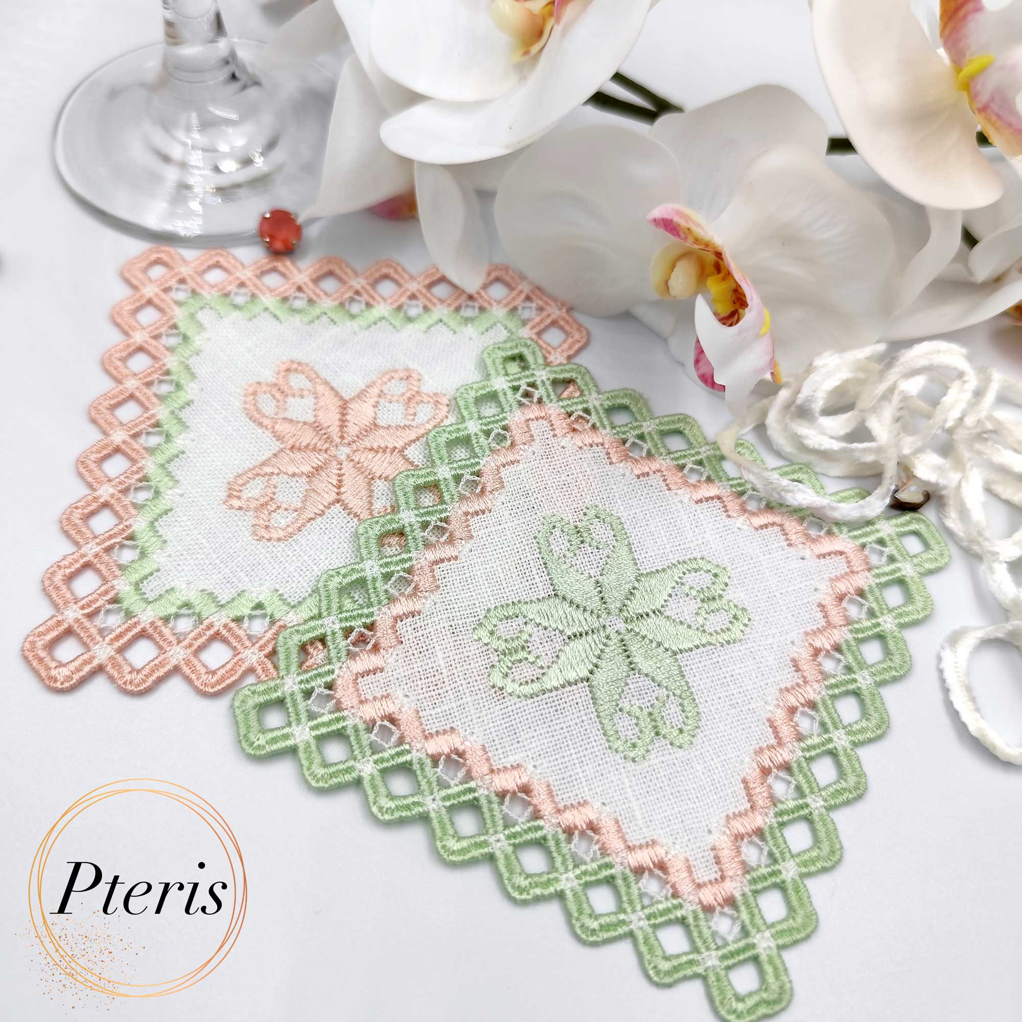 Square Hardanger cocktail napkin machine embroidery design displaying two linen napkins with a light green and peach embroidered geometric cutwork lace frame, styled flat next to white orchid blossoms.