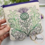 ITH quilted thistle makeup bag machine embroidery design showcasing a completed cream faux leather cosmetic pouch with a purple zipper and an intricately embroidered green and purple Scottish thistle, styled next to thread spools.