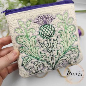 ITH quilted thistle makeup bag machine embroidery design showcasing a completed cream faux leather cosmetic pouch with a purple zipper and an intricately embroidered green and purple Scottish thistle, styled next to thread spools.