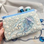 ITH quilted jellyfish makeup bag machine embroidery design showing a completed cream faux leather cosmetic pouch with a light blue zipper, featuring a detailed teal and purple embroidered jellyfish on a unique textured sea bubble quilted background.