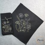 an elegant wild meadow grasses and wildflower botanical motif embroidered with white, cream, and green threads on a set of black linen napkins, styled on a textured grey background.