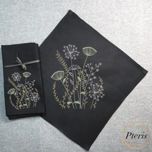 an elegant wild meadow grasses and wildflower botanical motif embroidered with white, cream, and green threads on a set of black linen napkins, styled on a textured grey background.