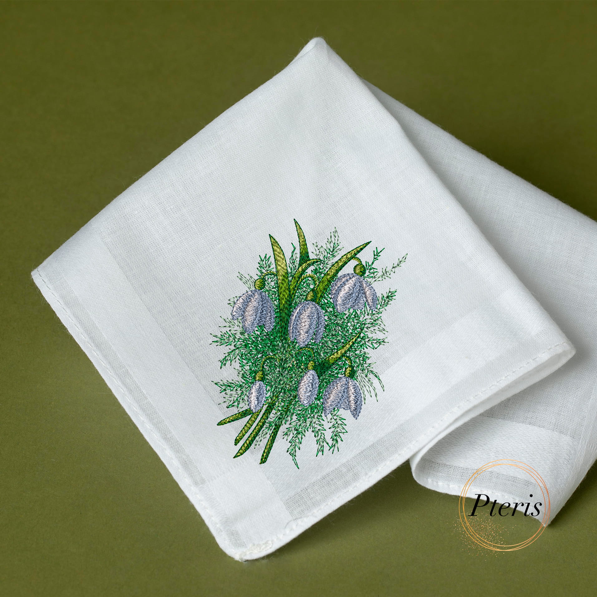 A delicate machine embroidery design of a spring flower bouquet with soft purple snowdrop blossoms and detailed green foliage, beautifully stitched on the corner of a folded white linen handkerchief, resting on a dark green surface, as shown in Easter-embroidery-design7.jpg.