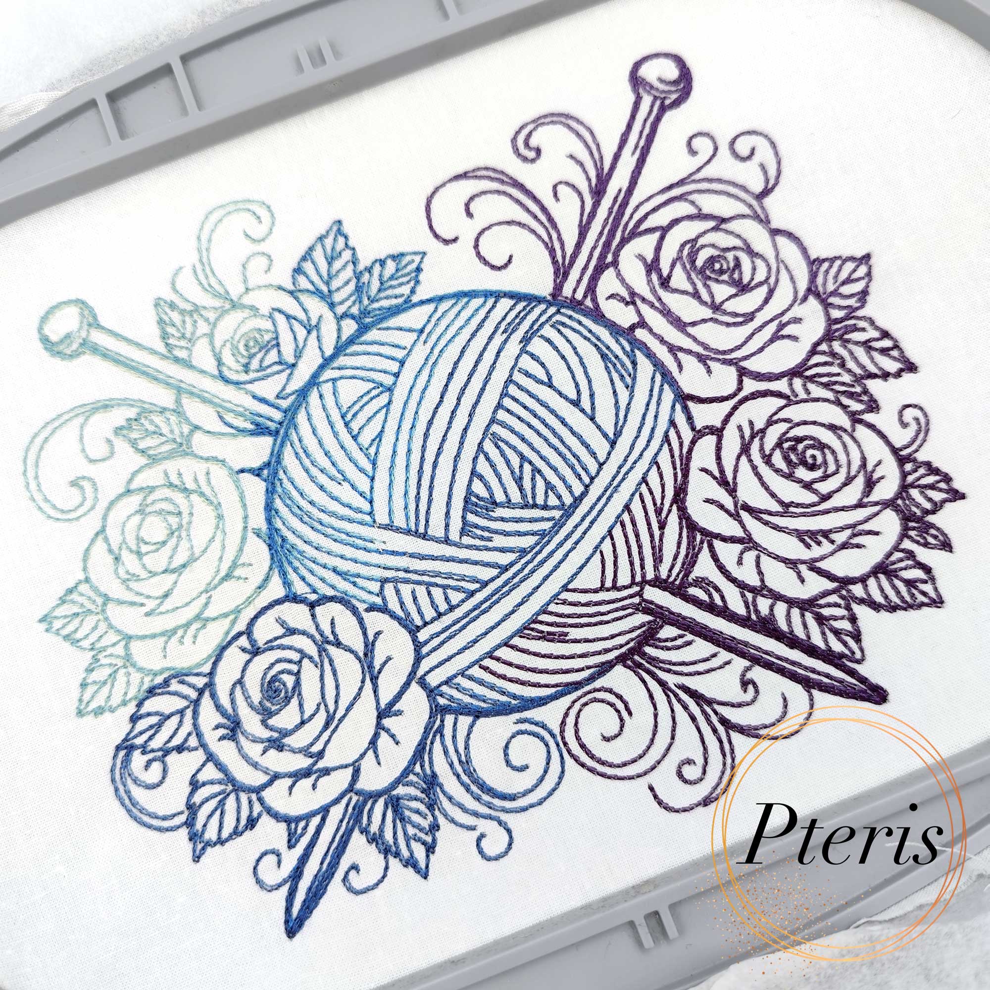 Machine embroidery design featuring a detailed ball of yarn with two knitting needles, surrounded by elegant roses and decorative swirls, stitched on white fabric using a blue-to-purple gradient thread.
