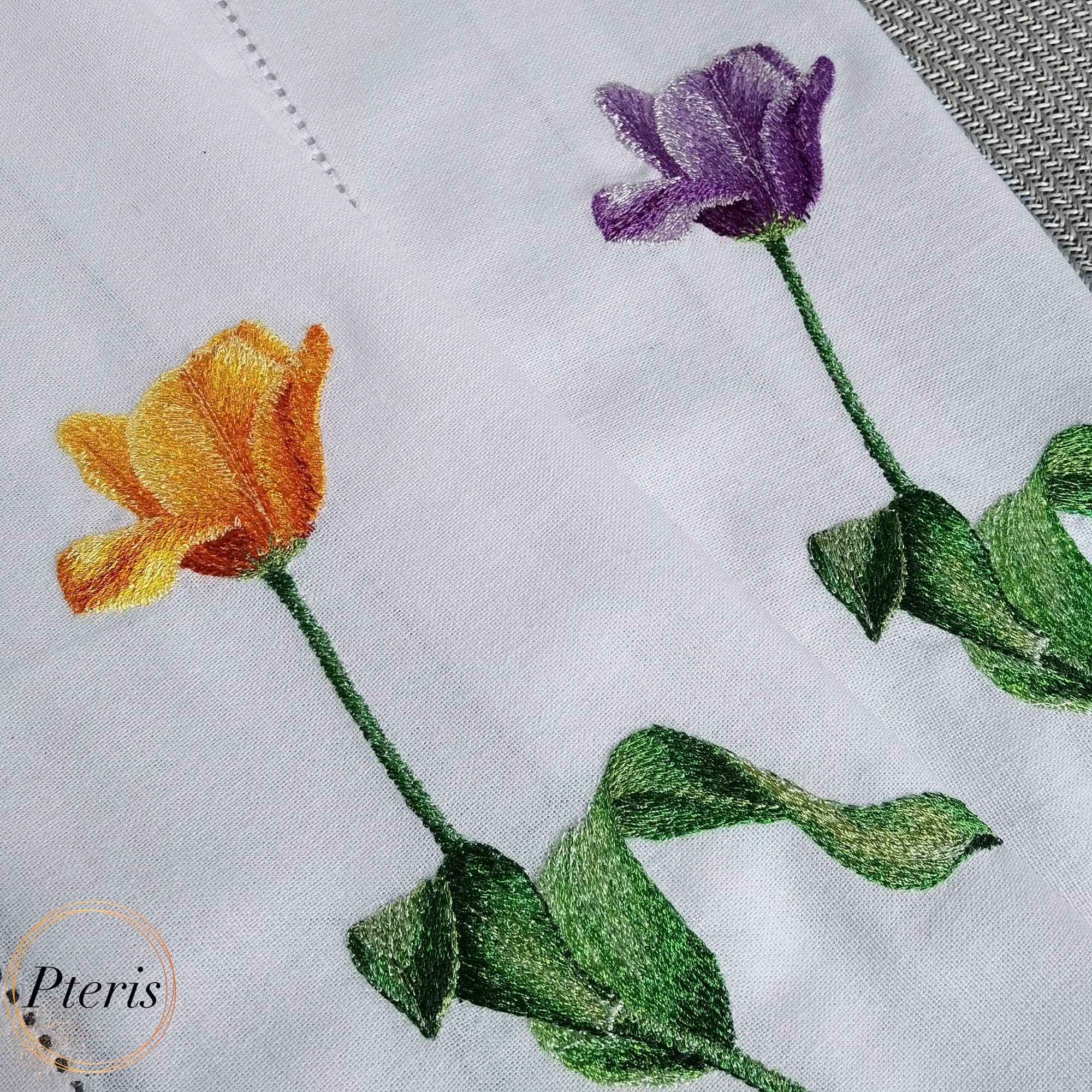 Close-up of machine-embroidered tulips in orange-yellow and purple colors with detailed shaded green leaves on a white linen towel.